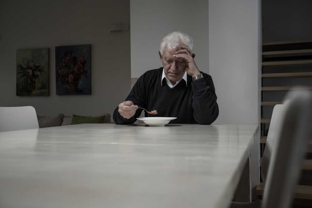 Senhor sentado a sua mesa de jantar de cabeça baixa, com a mão na testa, aparentando profunda tristeza.