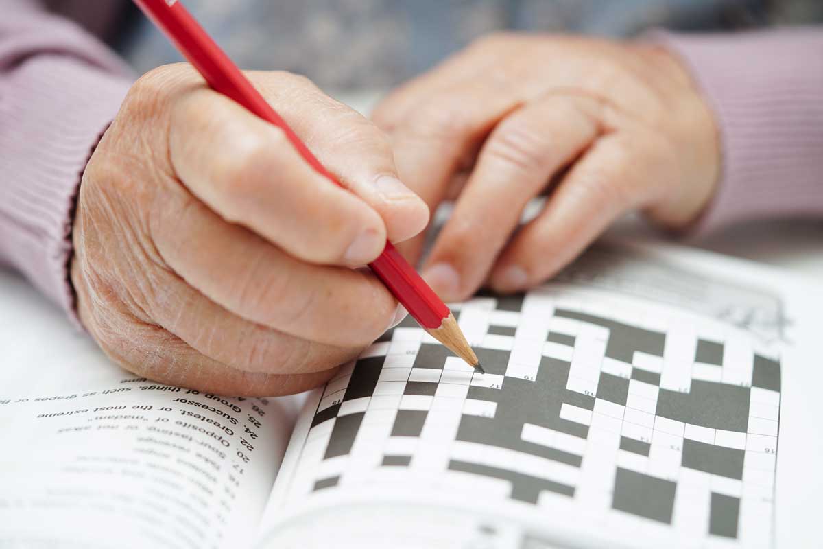 Mãos de pessoa idosa segurando um lápis e preenchendo um caderno de caça-palavras.