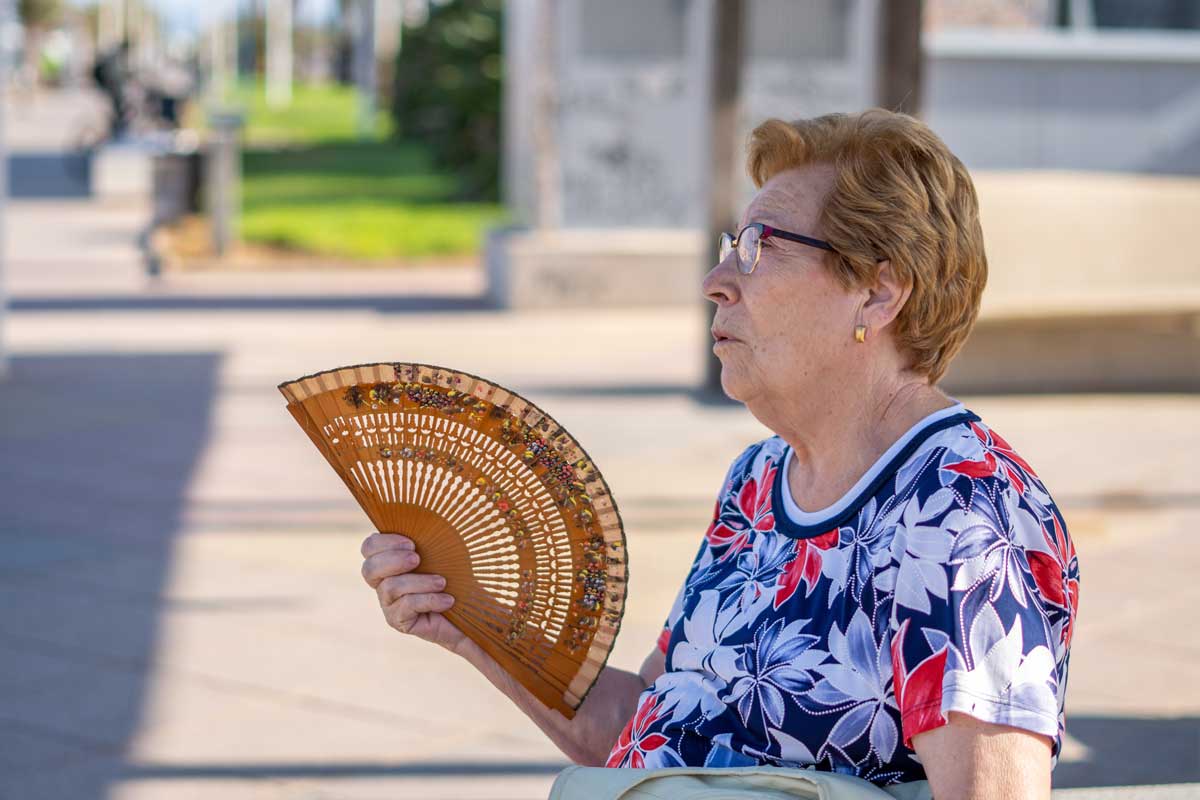 Pessoa idosa abanando um leque devido ao forte calor em dia ensolarado.